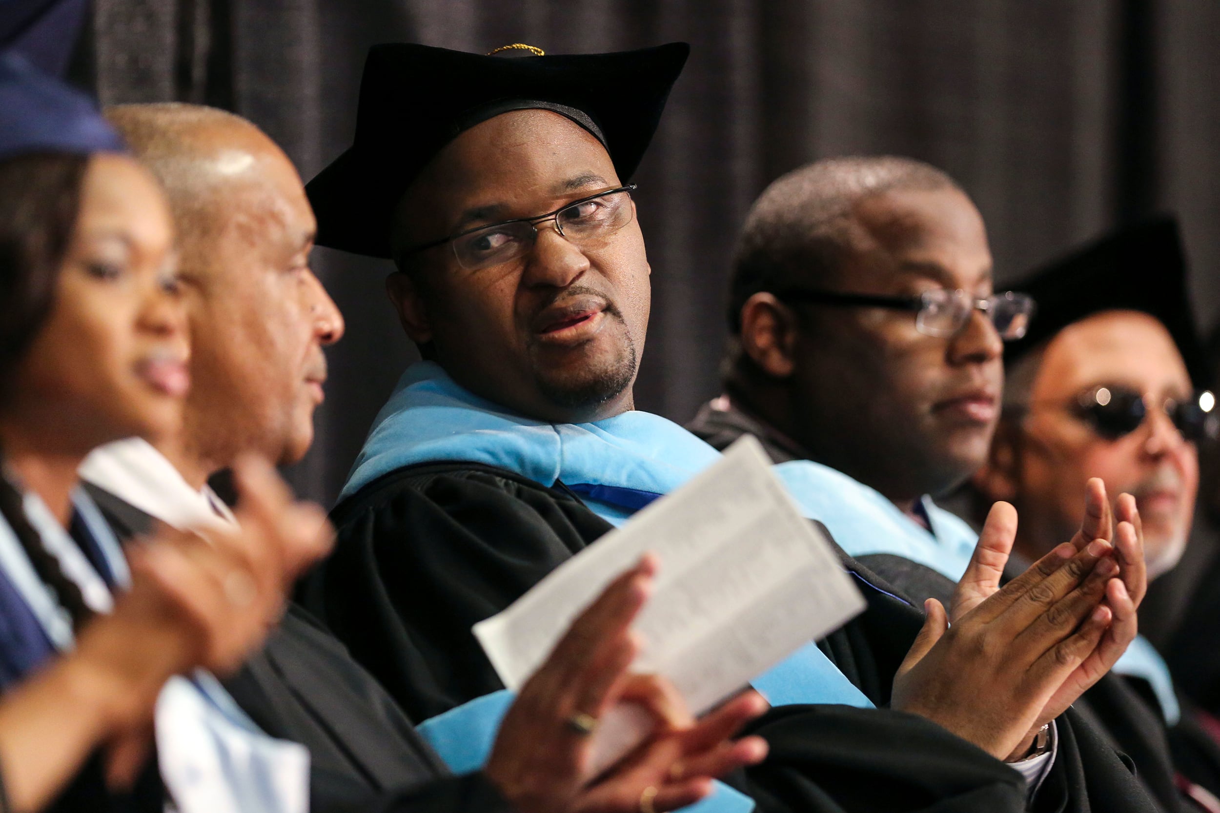 A photograph of adults wearing graduation caps and gowns sit during a ceremony talking with each other.