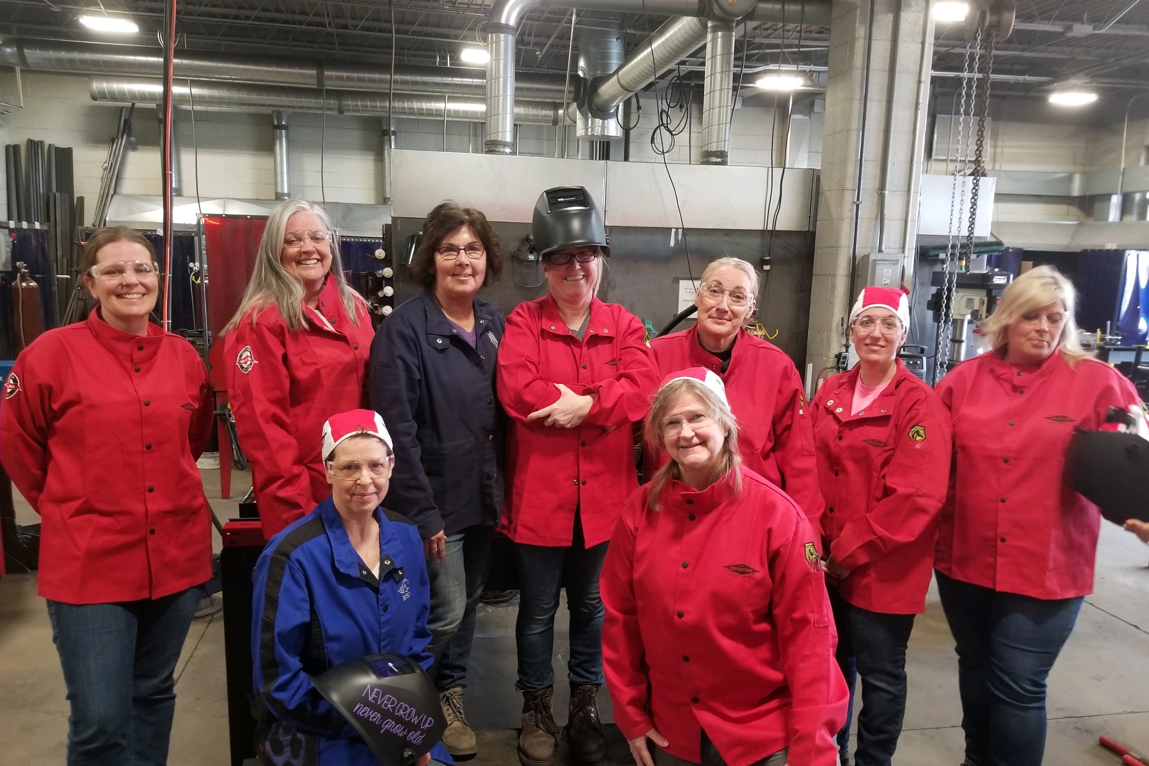 Nine women, mostly in red welding jackets, pose for a photo in a welding shop.