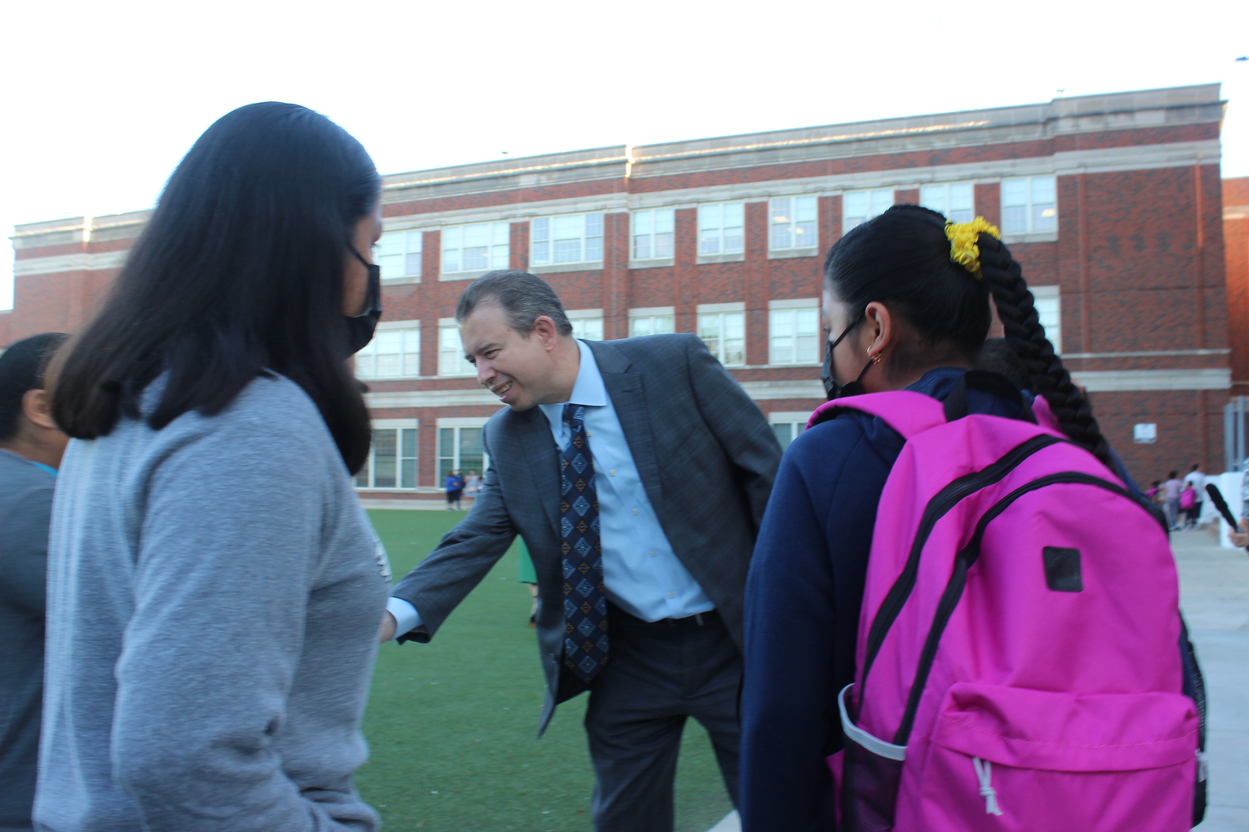 A man shakes hands with students wearing backpacks in front of a school building.