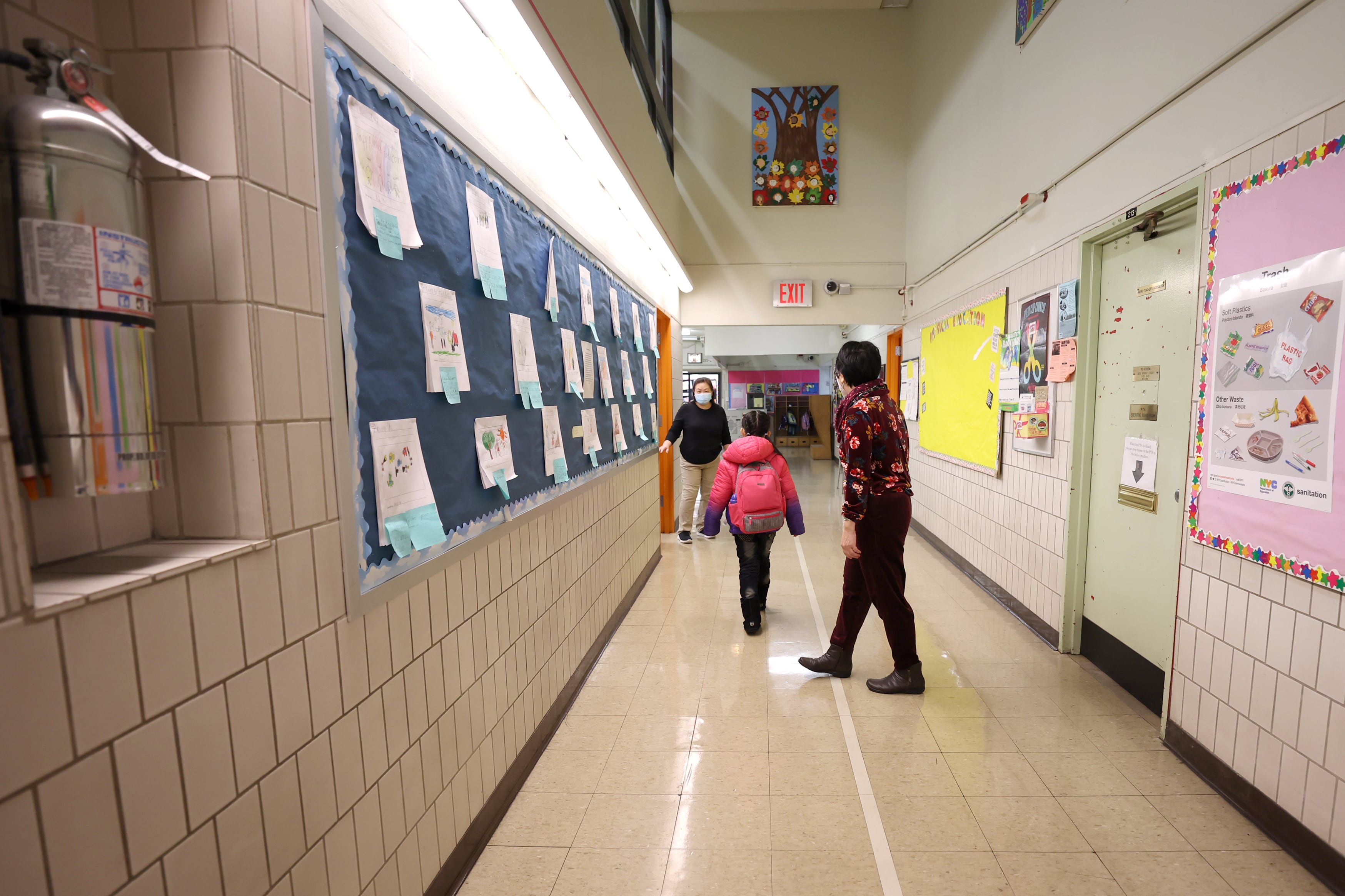 A principal walks a young student to her classroom in a school hallway. There are several pieces of student art and posters lining the tiled walls.