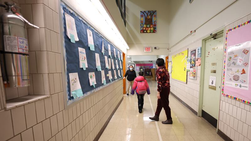 A principal walks a young student to her classroom in a school hallway. There are several pieces of student art and posters lining the tiled walls.