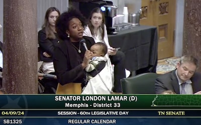 A screenshot of a woman holding a microphone and a young baby with three people in the background.