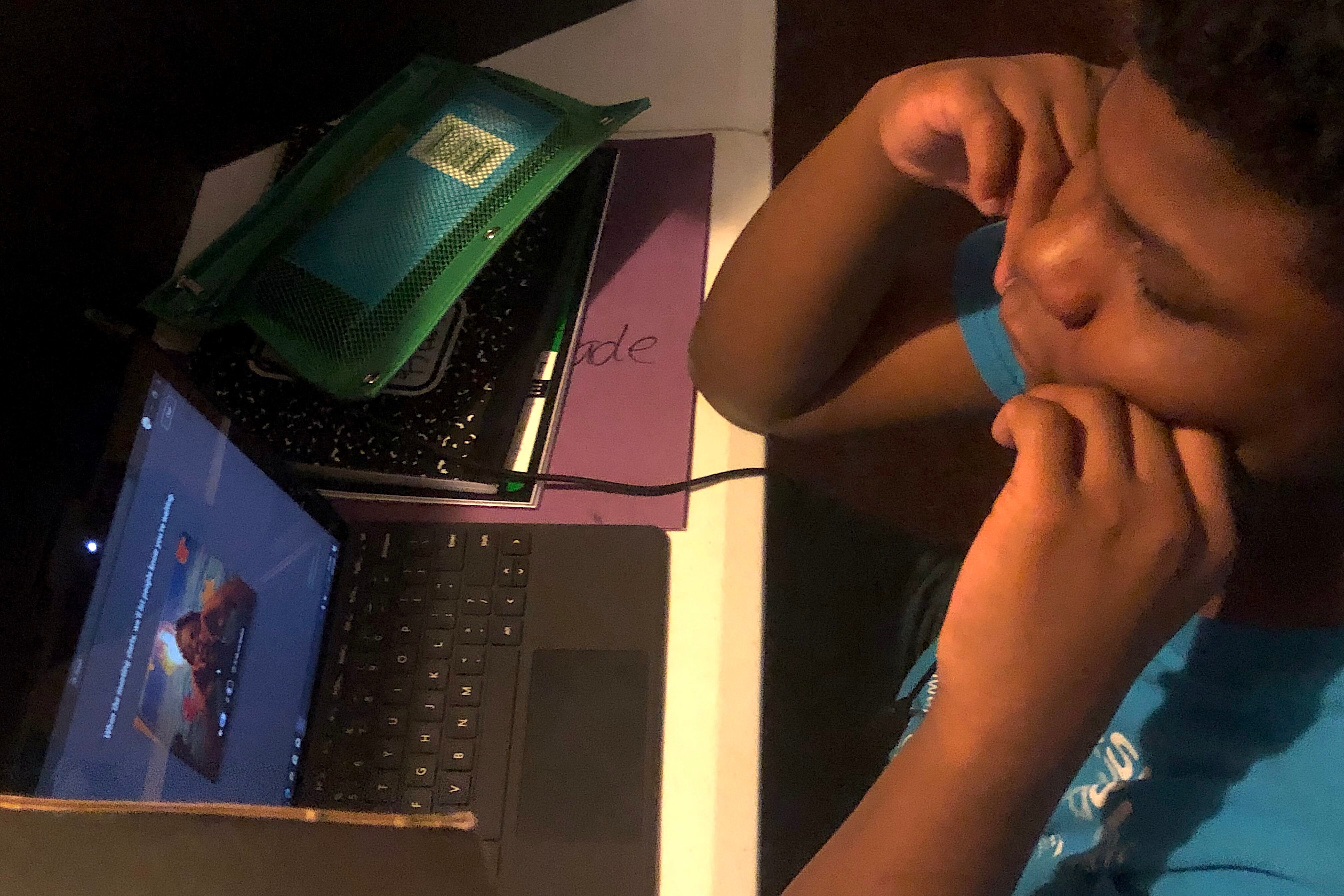 A student sits at a desk waiting for his online class via videoconference to start with a notebook and other school supplies nearby.