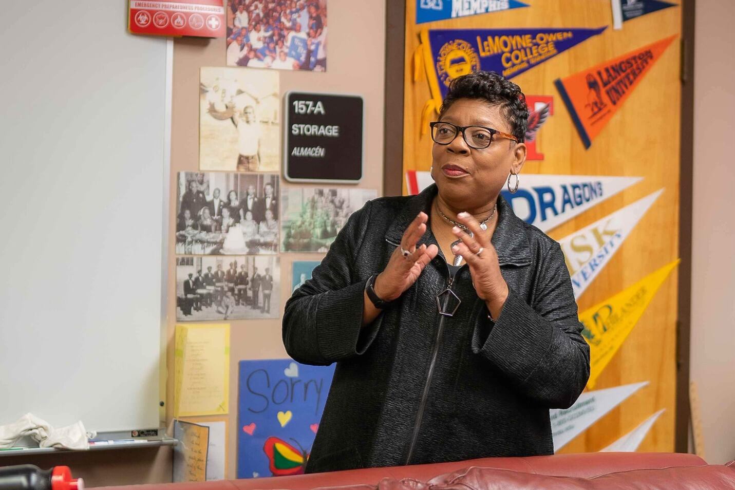 A woman wearing glasses speaks while gesturing with her hands in front of a door covered in college pendant flags