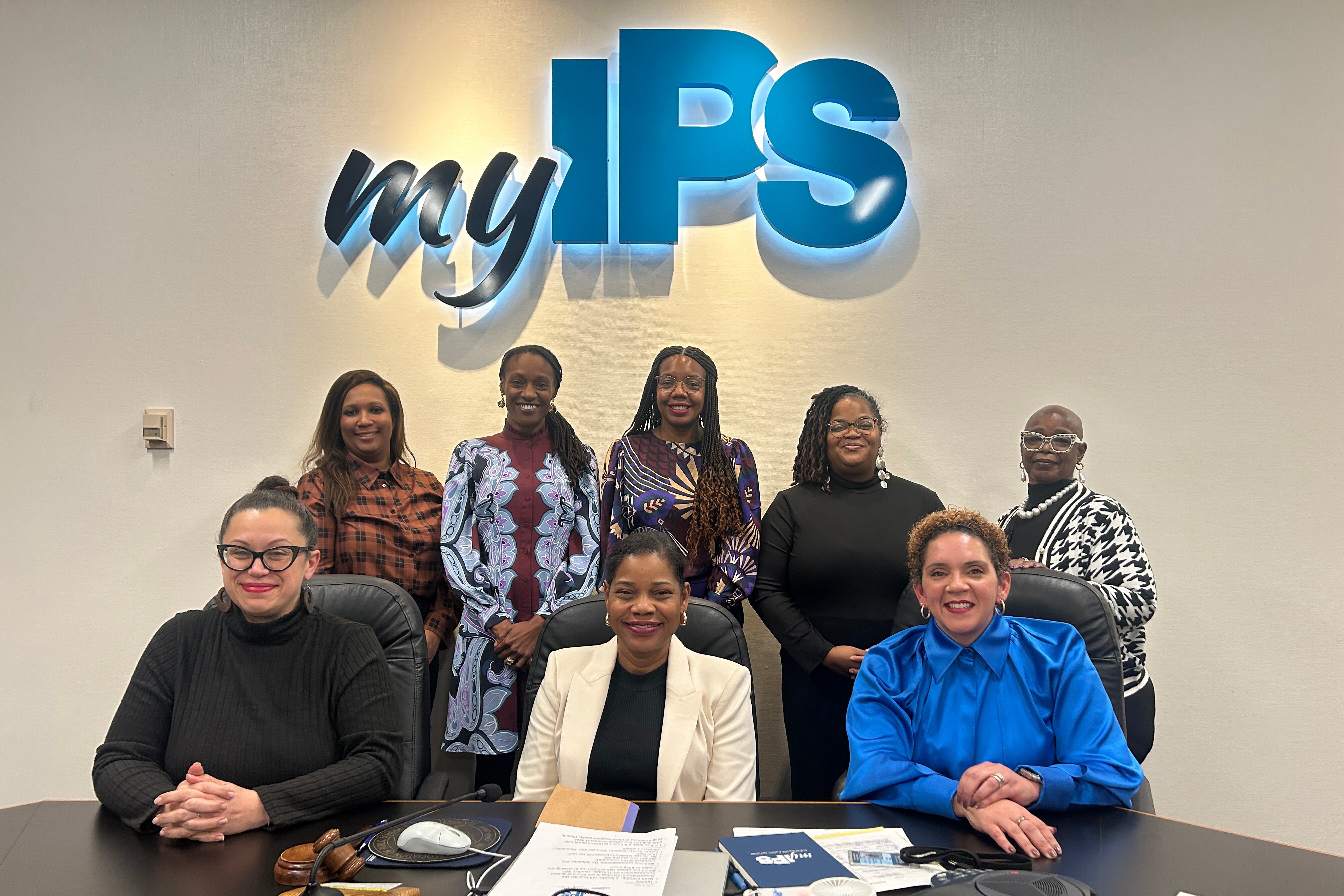 A photograph of eight women in business clothes pose for a portrait against a white wall with an illuminated sign that reads "My IPS."
