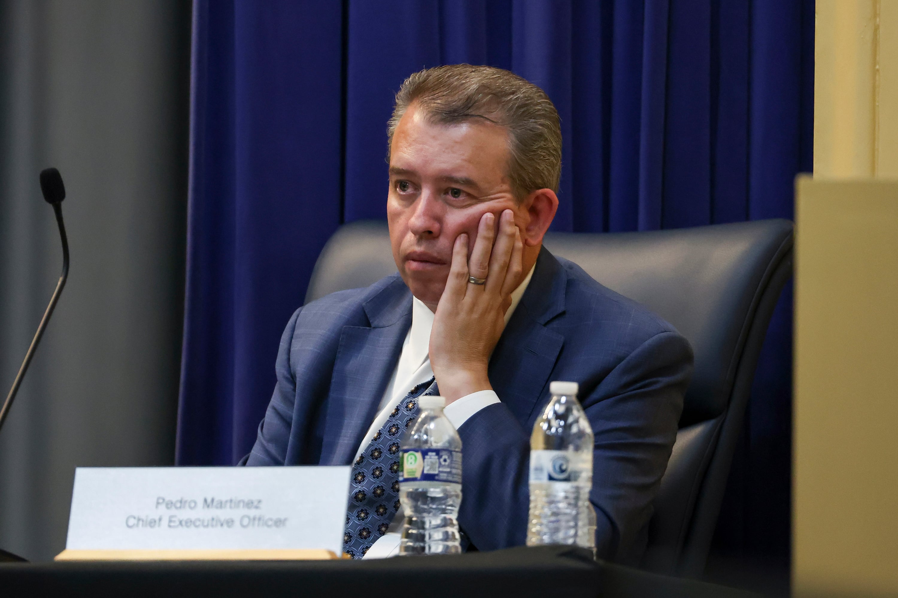 A man in a suit sits behind a microphone with two bottles of water on the table next to a name card.