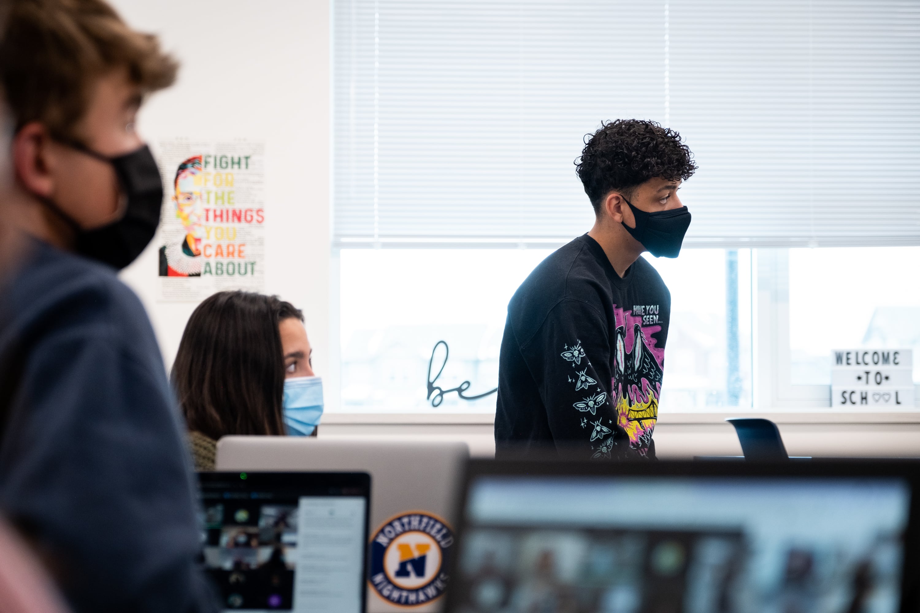 A high school student sits on top of a desk in a classroom. He’s in profile. He’s wearing a black face mask. Other students have masks too.