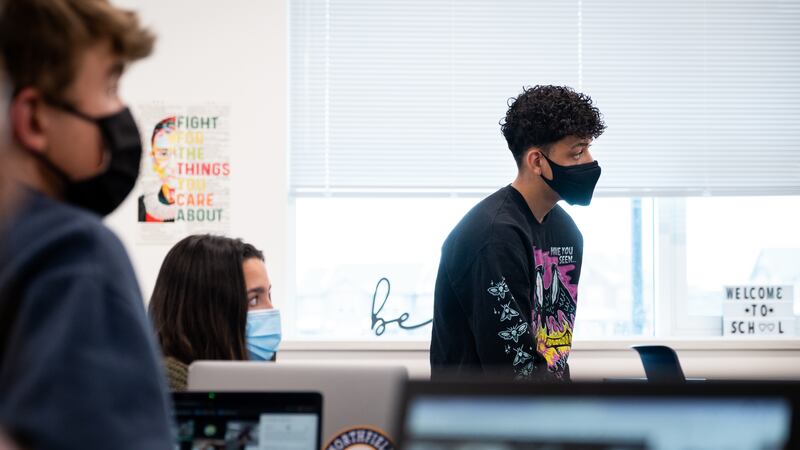 A high school student sits on top of a desk in a classroom. He’s in profile. He’s wearing a black face mask. Other students have masks too.