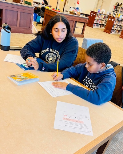 A female tutor wearing a blue college sweatshirt sits at a table in the library working with a young student on a math assignment.