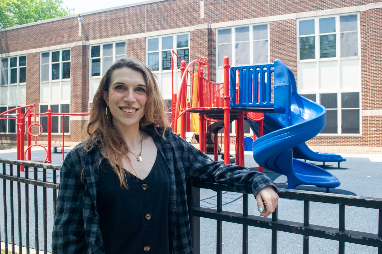 A woman in a flannel and tank top rests her arm on the railing of a black metal fence, which reaches up to her chest. Her face is to the camera and she is smiling. She has light brown hair that reaches a bit past her shoulders. She has a septum piercing and is wearing a delicate gold necklace. Behind the fence is playground equipment, including a curved slide. Behind the playground is a brick elementary school building.