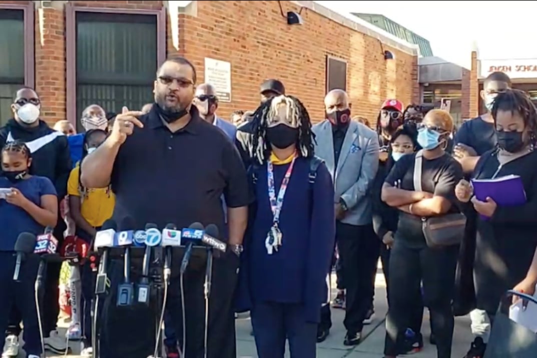Shauntee Colston, a parent at Jensen Elementary on Chicago’s West Side, stands behind a stand of microphones, outside the school building with his daughter, who is wearing a face mask, and other parents.