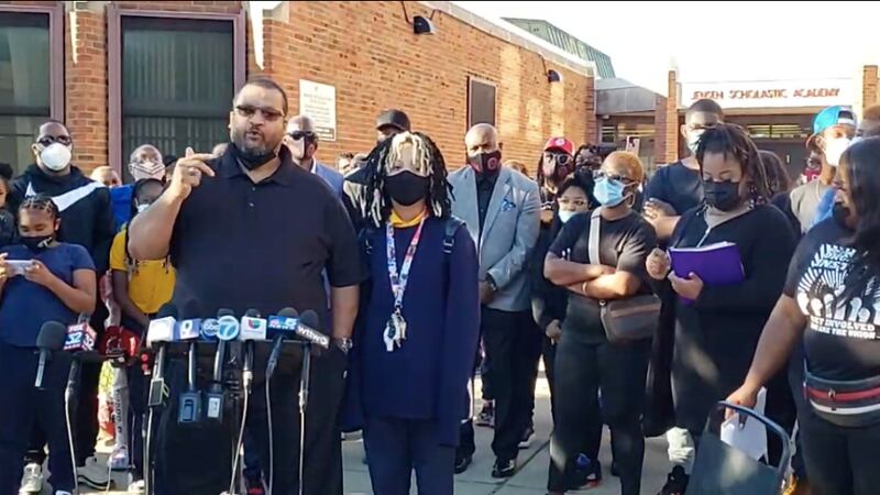 Shauntee Colston, a parent at Jensen Elementary on Chicago’s West Side, stands behind a stand of microphones, outside the school building with his daughter, who is wearing a face mask, and other parents.