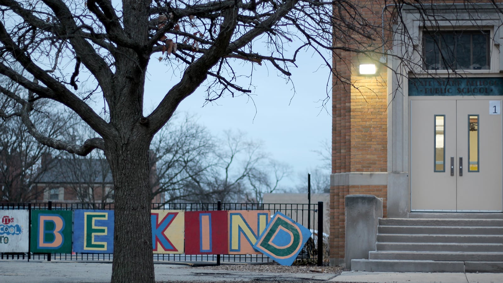 A sign that reads "Be Kind" is on display on a fence next to an entrance of a school with a tree in the foreground.