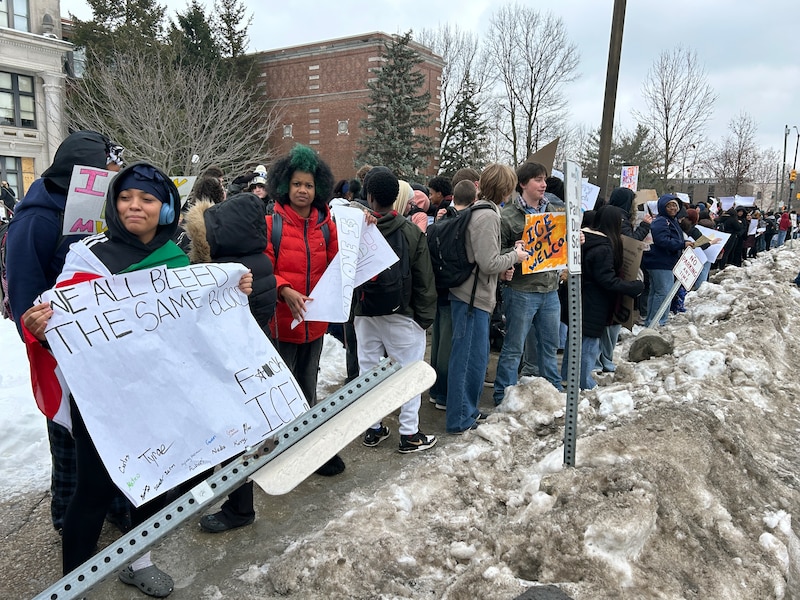 A photograph of a large group of high school students, some holding protest signs and all dressed in coats and cold weather gear stand outside on a cold, cloudy day protesting against ICE.