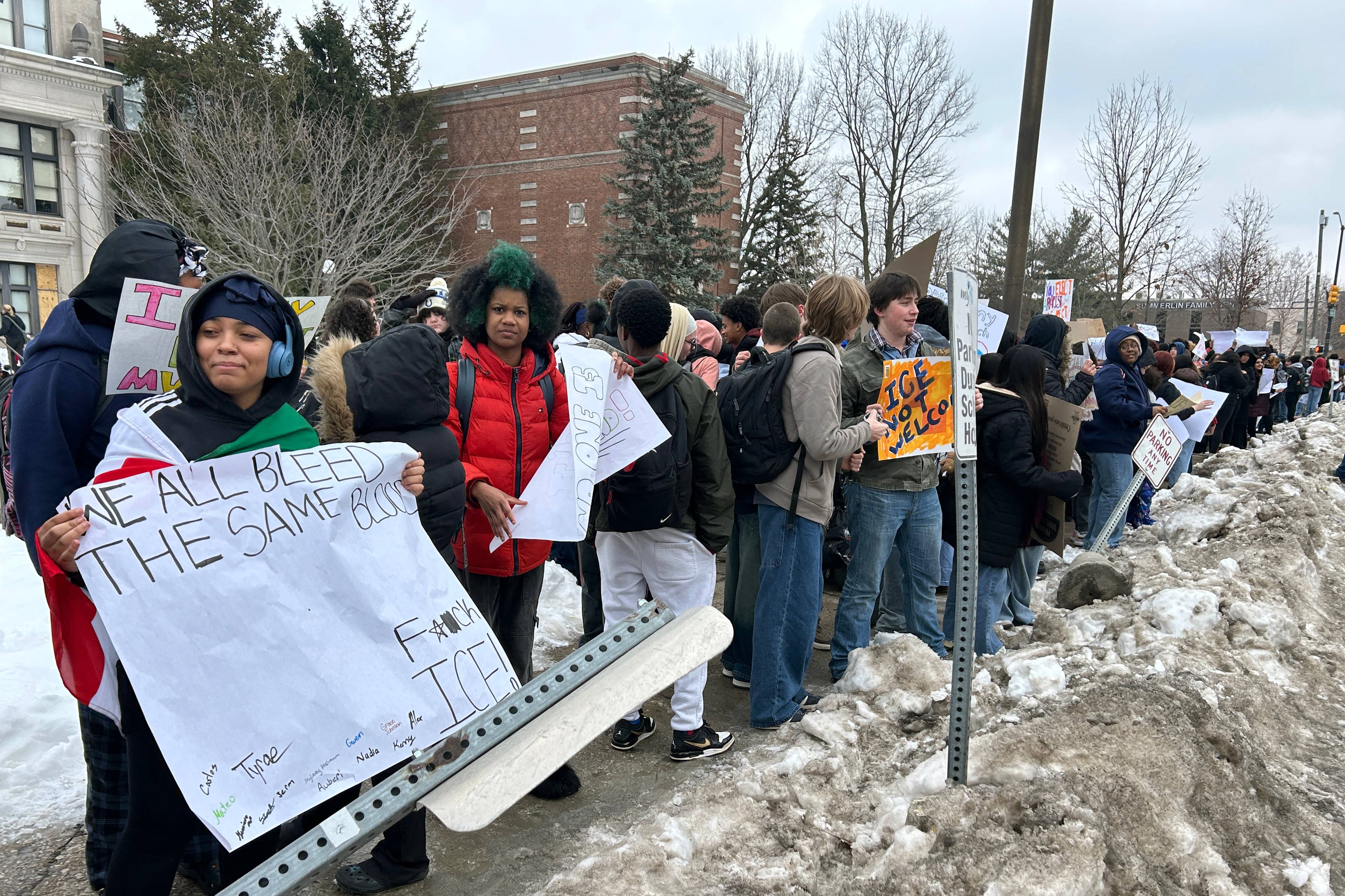 A photograph of a large group of high school students, some holding protest signs and all dressed in coats and cold weather gear stand outside on a cold, cloudy day protesting against ICE.