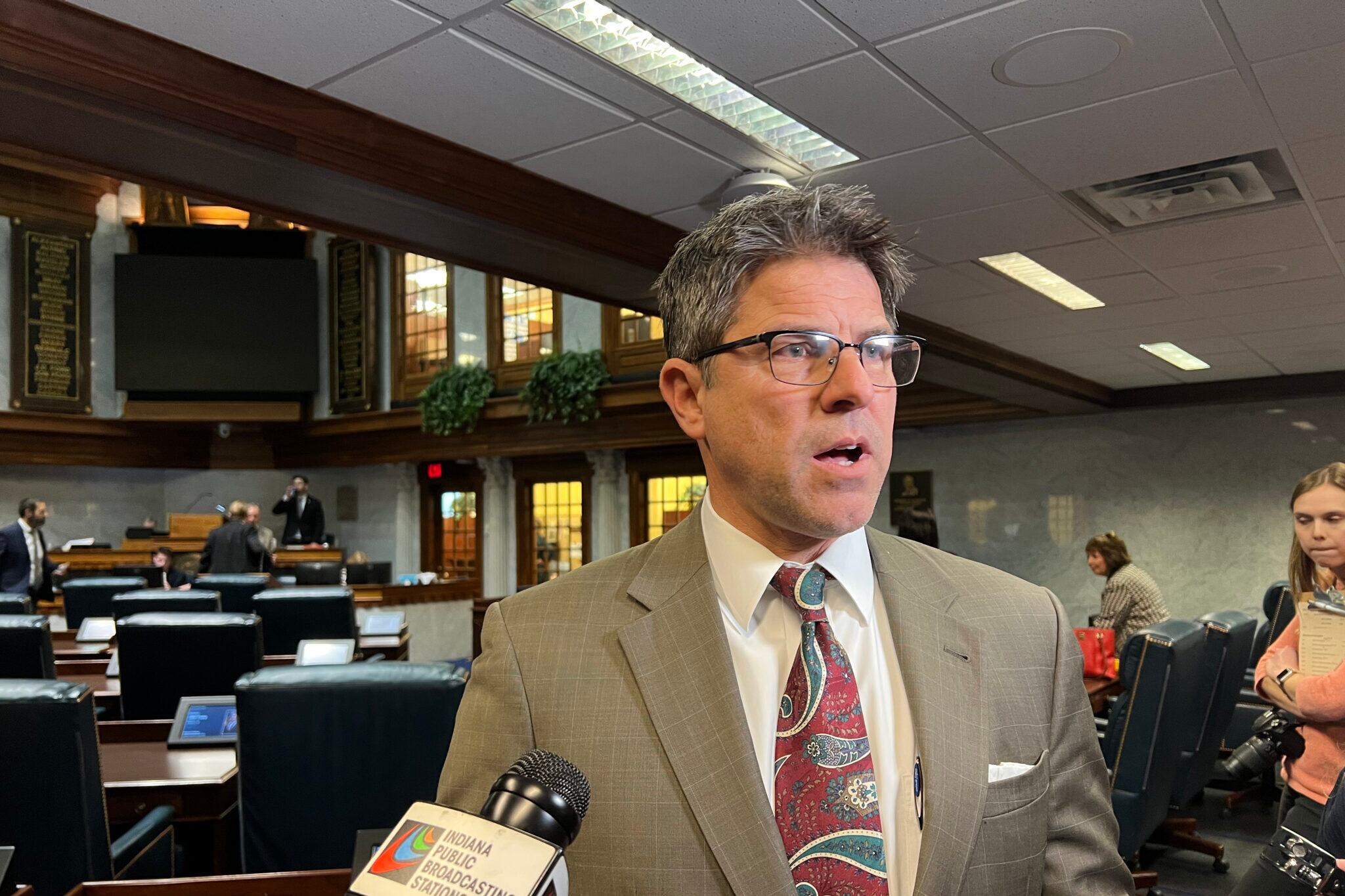 A man wearing glasses, a red tie, and tan suit speaks in front of microphones