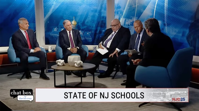 Five men in suits sit on chairs on a TV news set.
