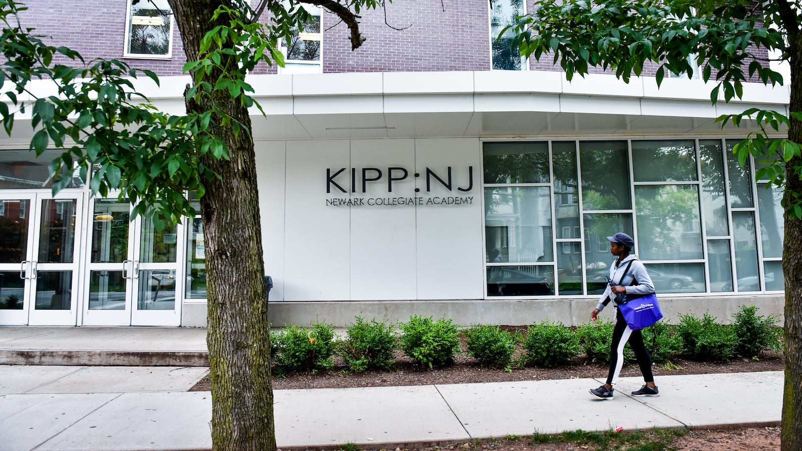 A student wearing a cap and carrying a blue tote bag walks on a walkway in front of the KIPP-NJ building.