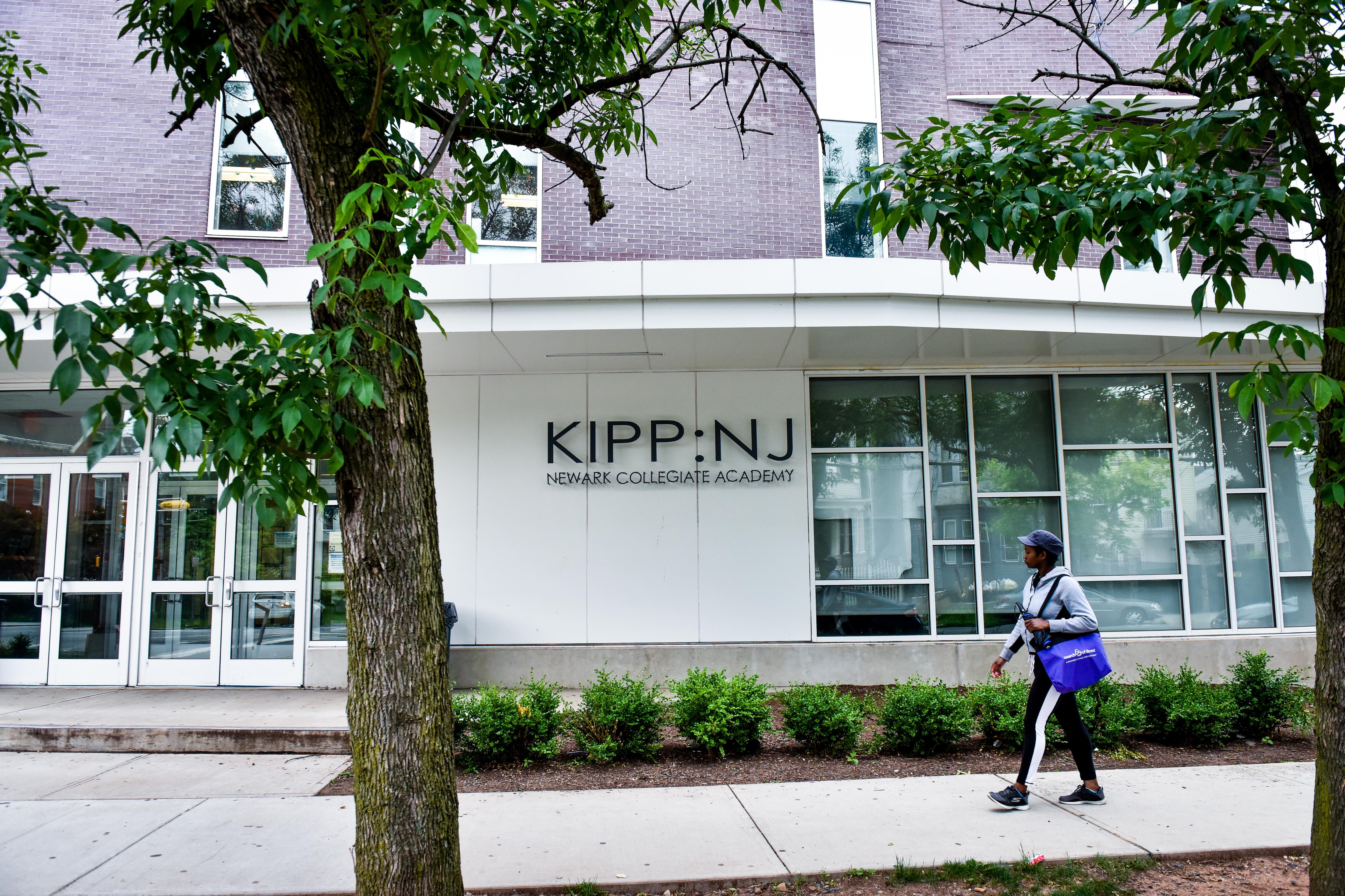 A student wearing a cap and carrying a blue tote bag walks on a walkway in front of the KIPP-NJ building.