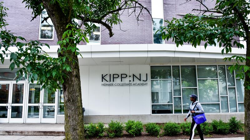 A student wearing a cap and carrying a blue tote bag walks on a walkway in front of the KIPP-NJ building.