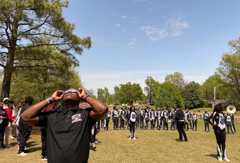 A group of high school students in the marching band perform while one student wears eclipse viewing glasses outside with green grass and a clear blue sky.