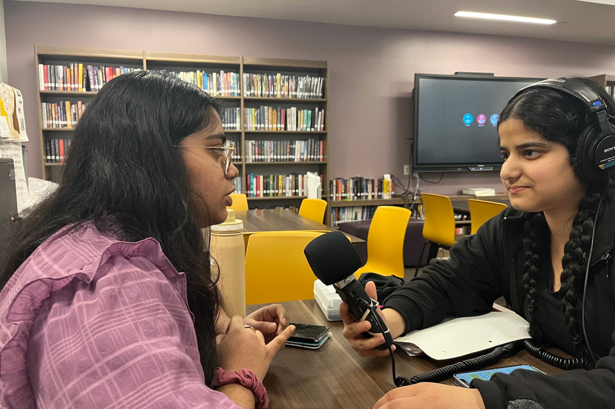 Two high school students sit at a table, the student on the left is wearing a pink jacket and the student on the right is wearing a black jacket and holding a microphone and wearing headphones.