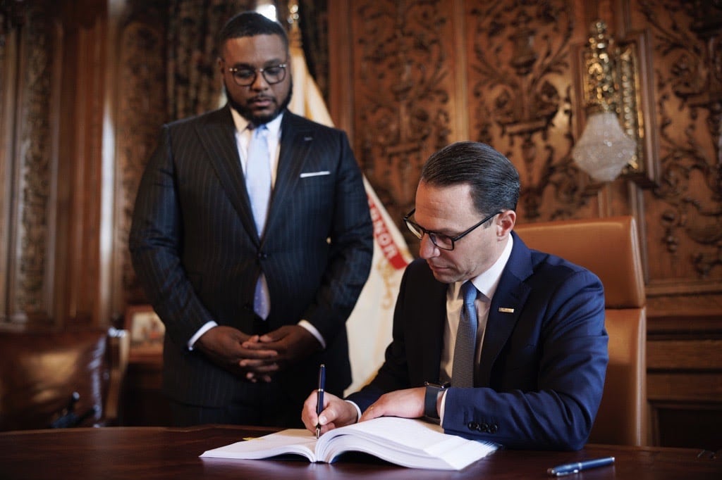 A man in a blue suit and blue tie sits at a table and signs a document as a man in a dark suit to his right looks on.