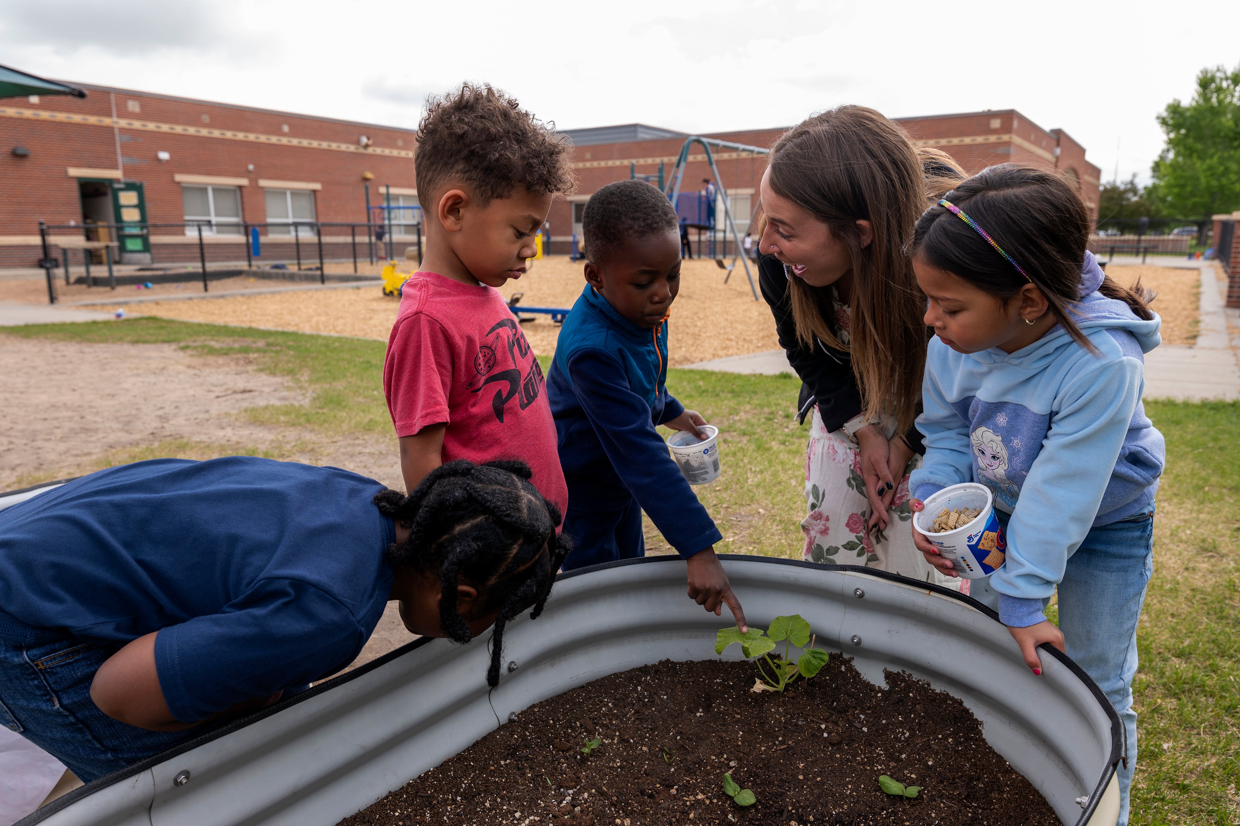 A preschool teacher leans over garden planter as four children look on.