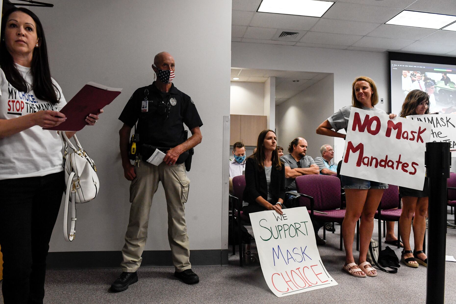 A security officer in a brown and biege uniform stands against a wall surrounded by women carrying signs that say “No mask mandates” and “Support mask choice.”
