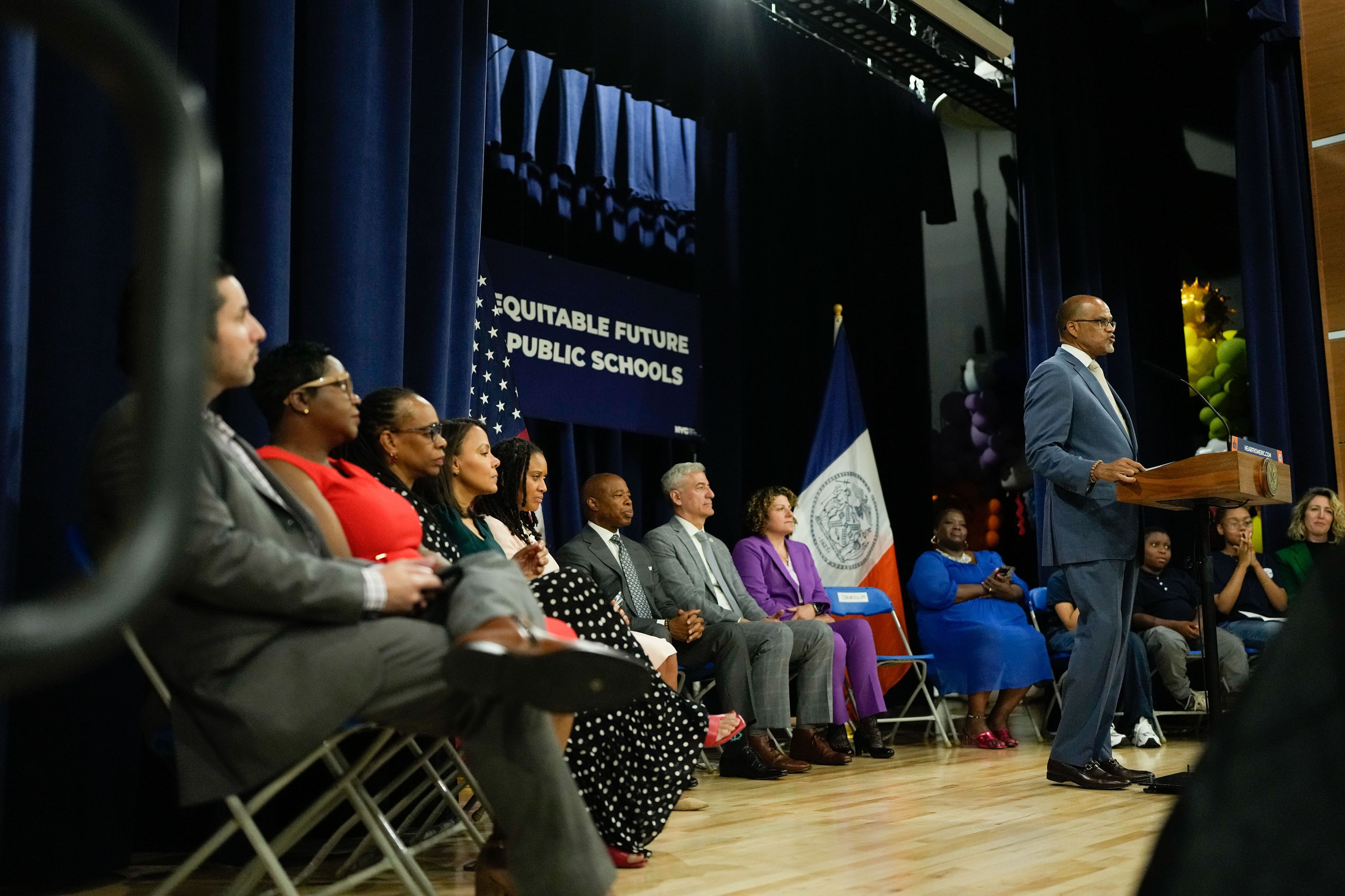 A man on the far right of the image wearing a blue suit stands behind a podium while a row of people sit in chairs in the background.