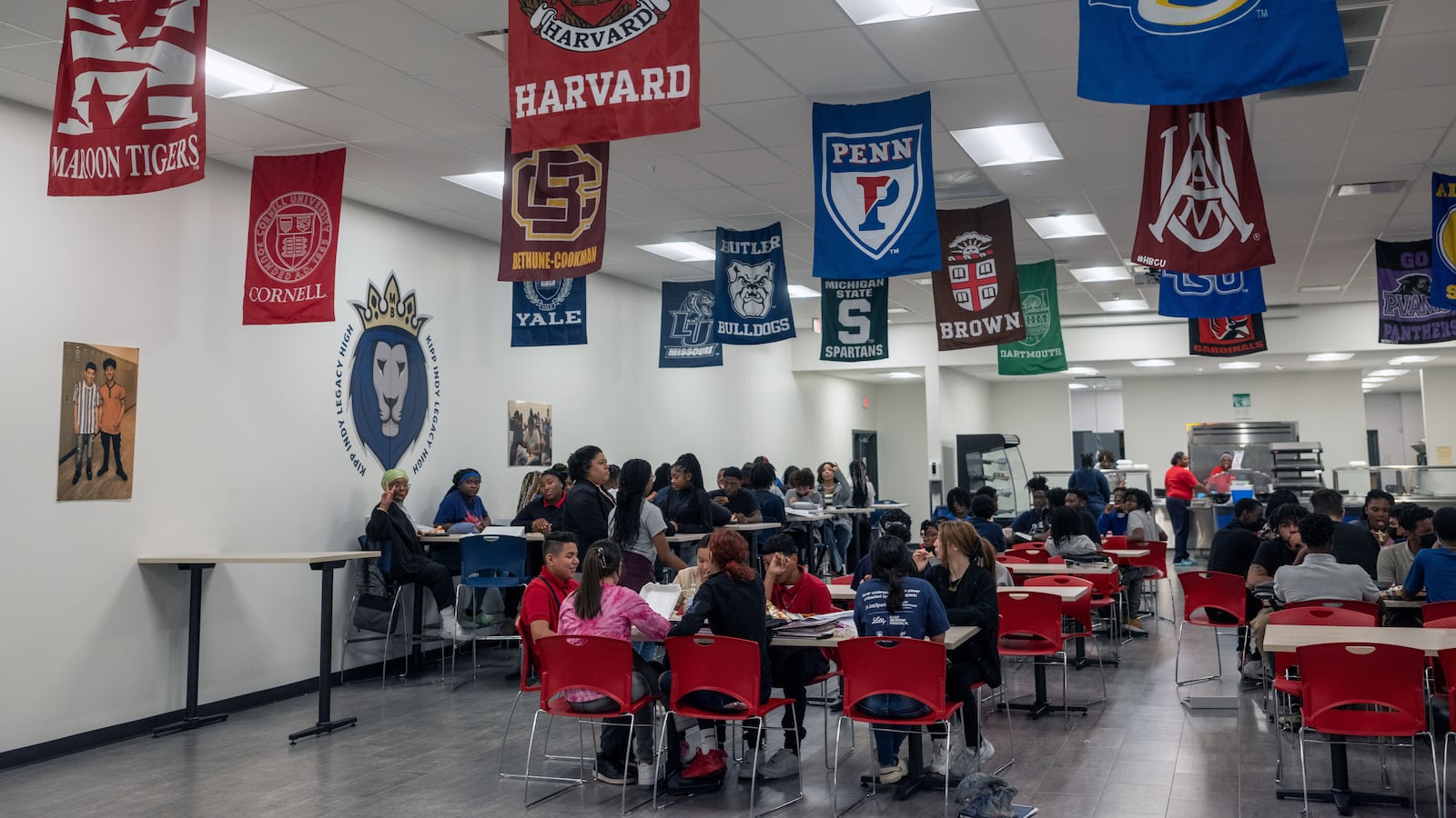 Flags of various colleges hang from the ceiling of a high school cafeteria as students sit at their tables.