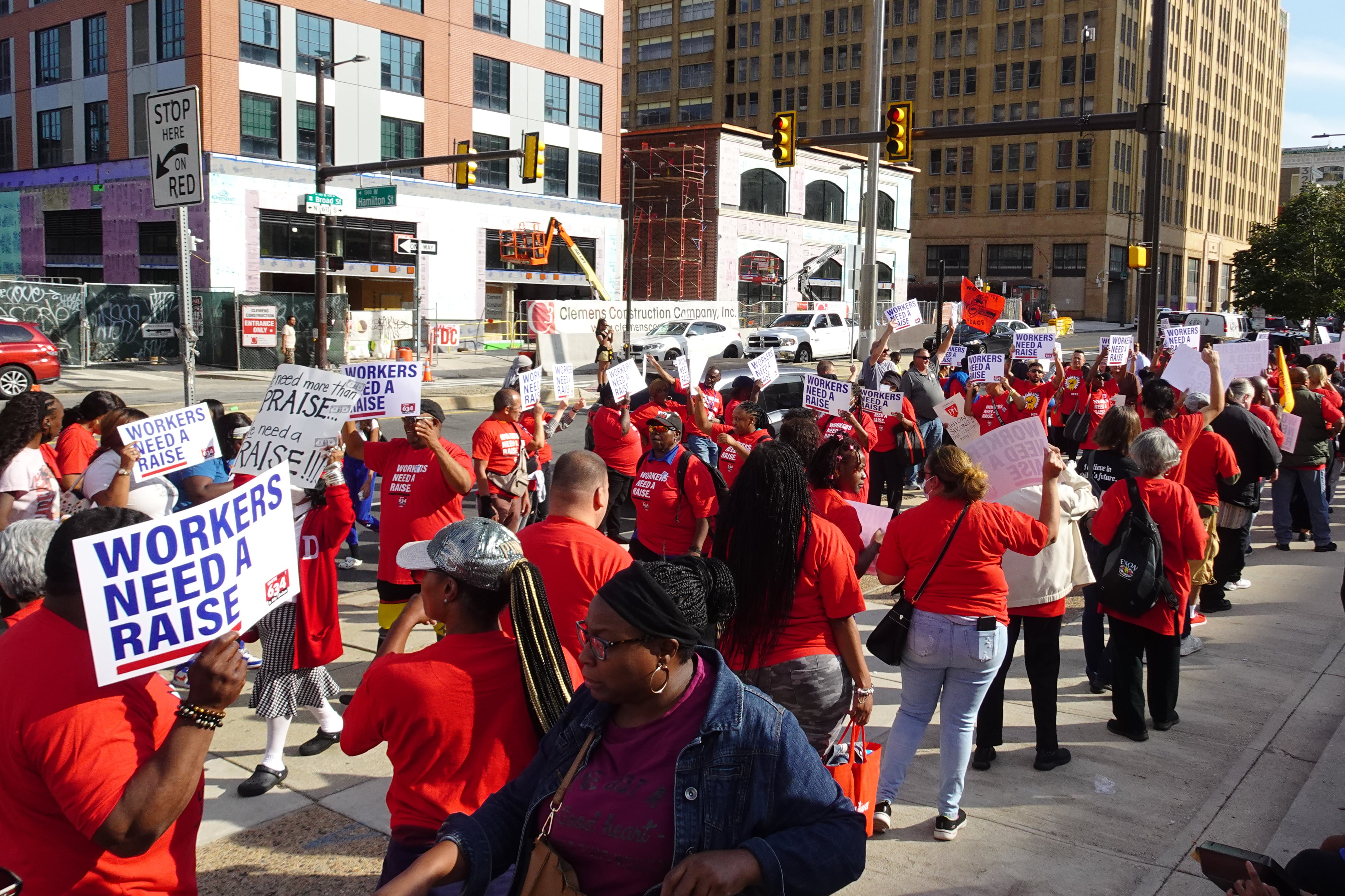 A group of people in red shirts march carrying signs that say “workers need a raise.”