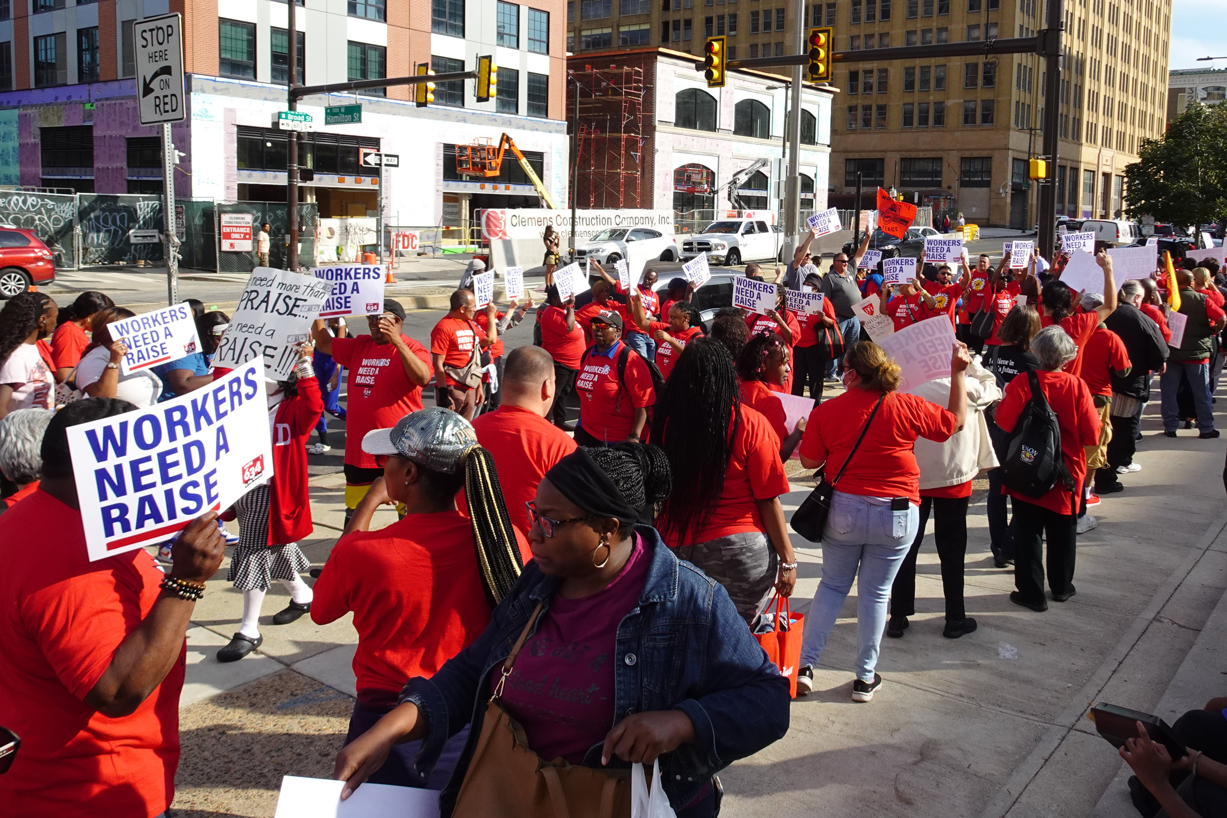 A group of people in red shirts march carrying signs that say “workers need a raise.”