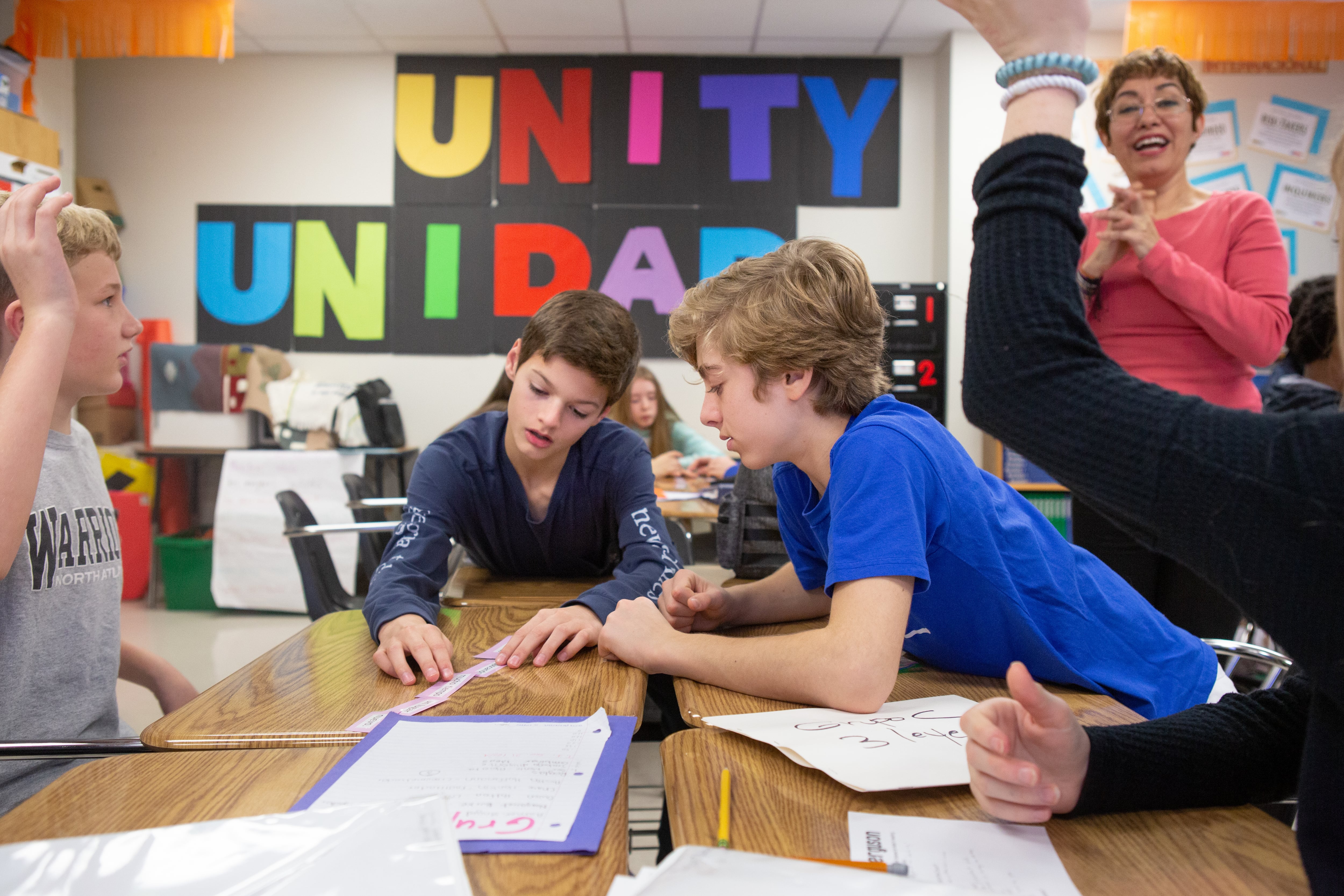 Young students work on their classwork in groups while the teacher stands behind them, clapping their hands in support. The wall in the background has colorful letters that read “Unity, Unidad”