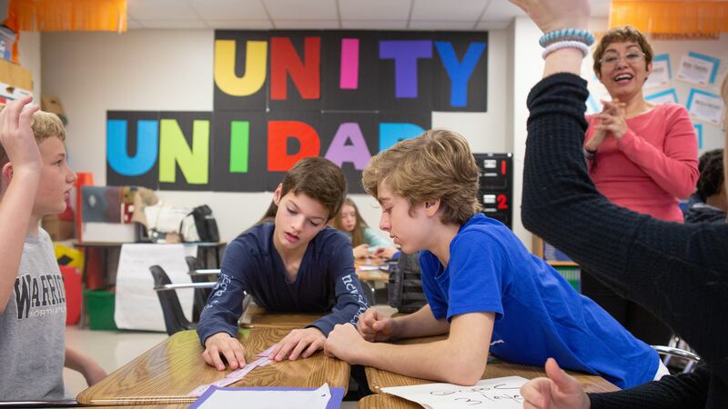 Young students work on their classwork in groups while the teacher stands behind them, clapping their hands in support. The wall in the background has colorful letters that read “Unity, Unidad”