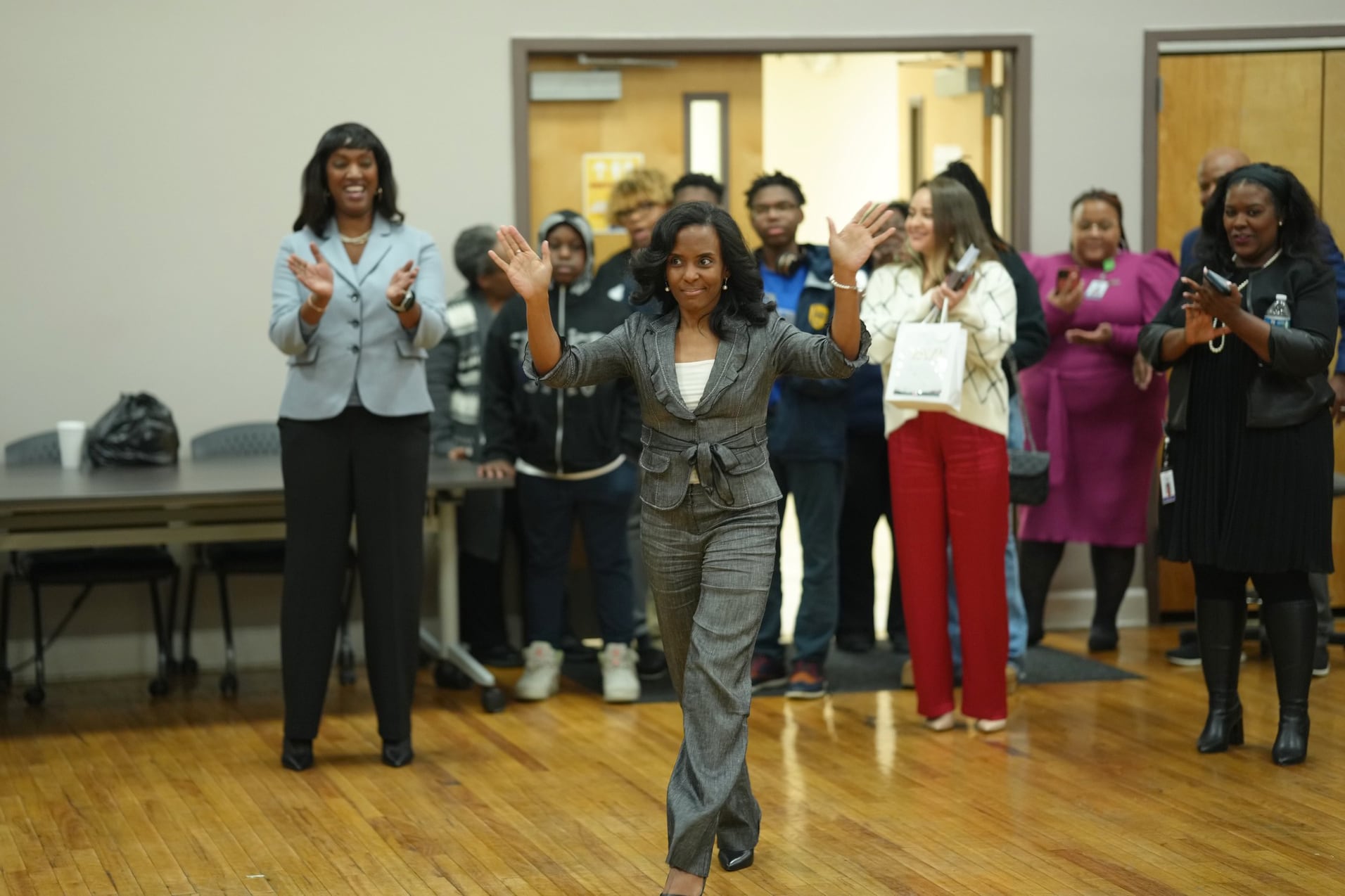 A woman wearing a dark suit jacket waves and walks in front of a crowd of people in the background.