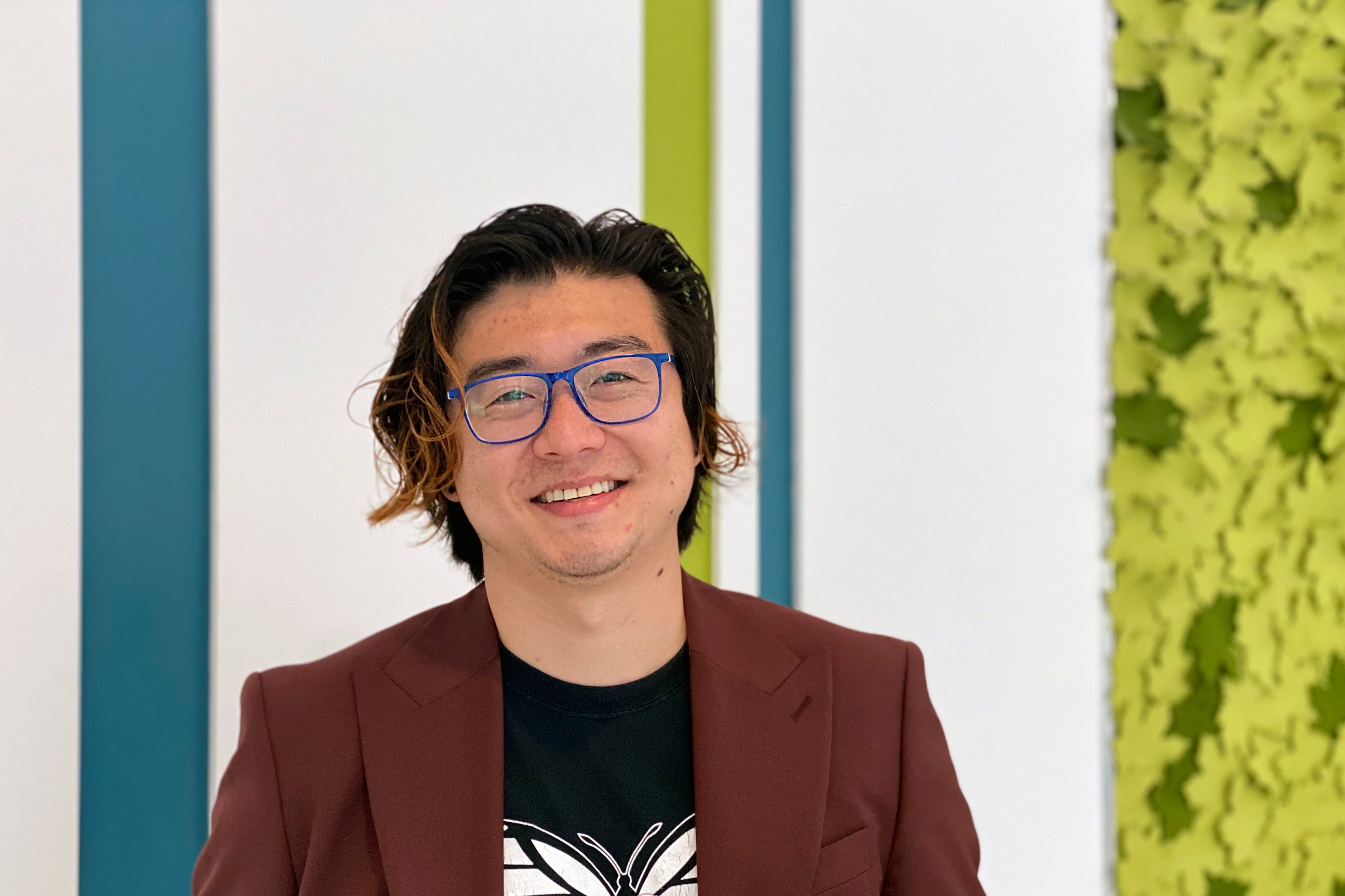 A man with glasses and wearing a T-shirt with a butterfly under a blazer looks at the camera.