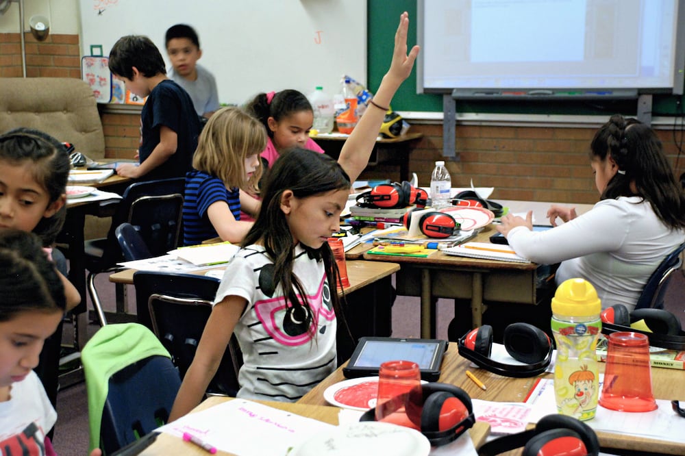 A little girl raises her hand. She is sitting at a desk in a classroom, surrounded by other students.