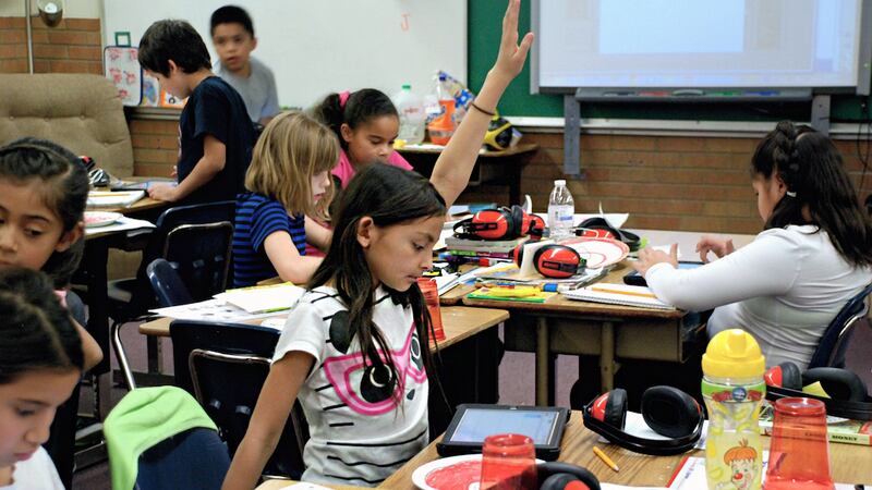 A little girl raises her hand. She is sitting at a desk in a classroom, surrounded by other students.