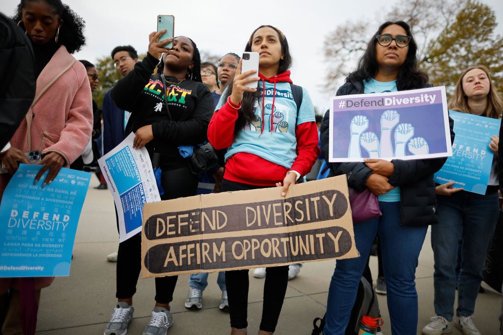 A group of young people stand in a group on a gray day. They are holding signs in support of affirmative action and diversity.