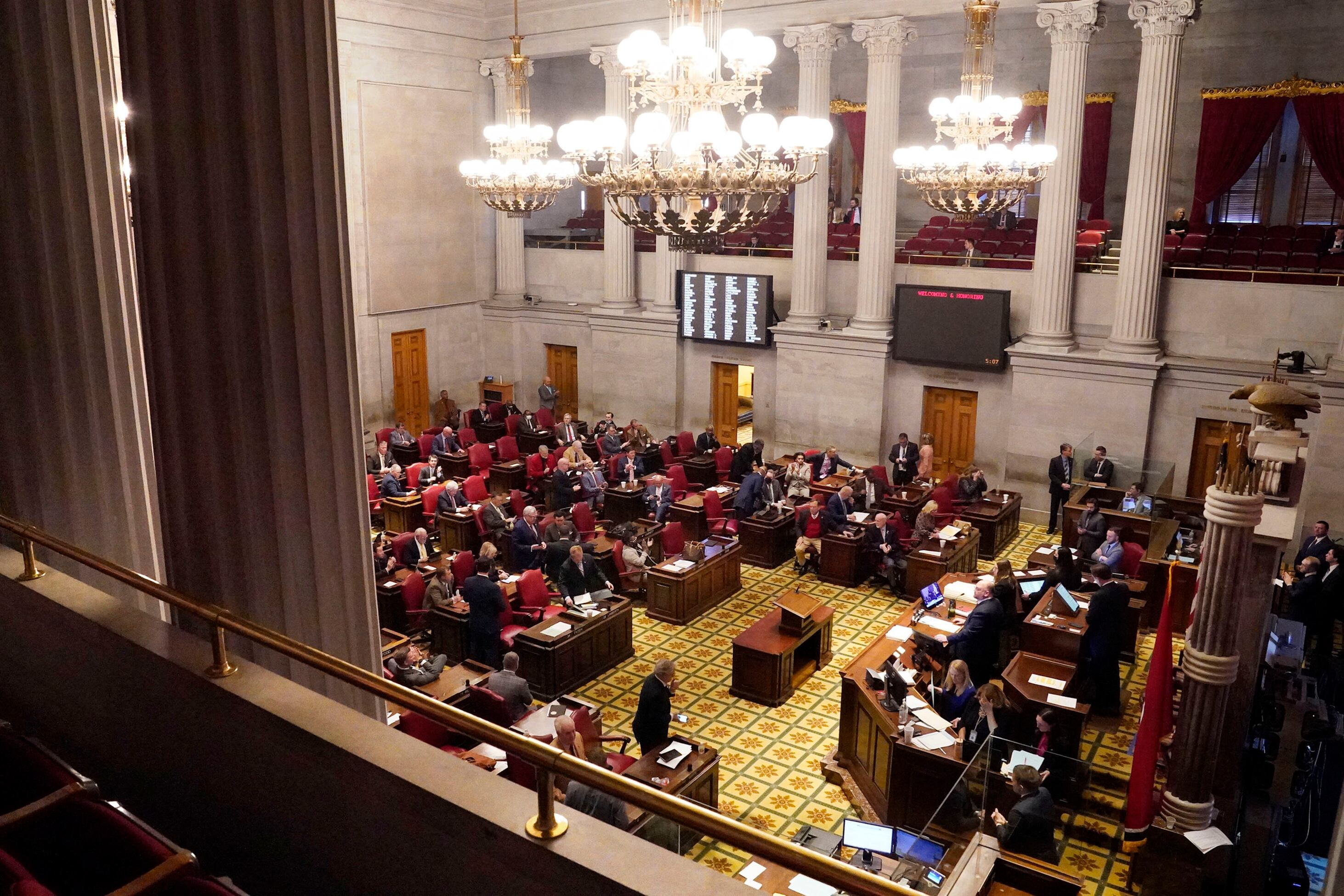 Dozens of people sit at desks or mill around a large legislative chamber.