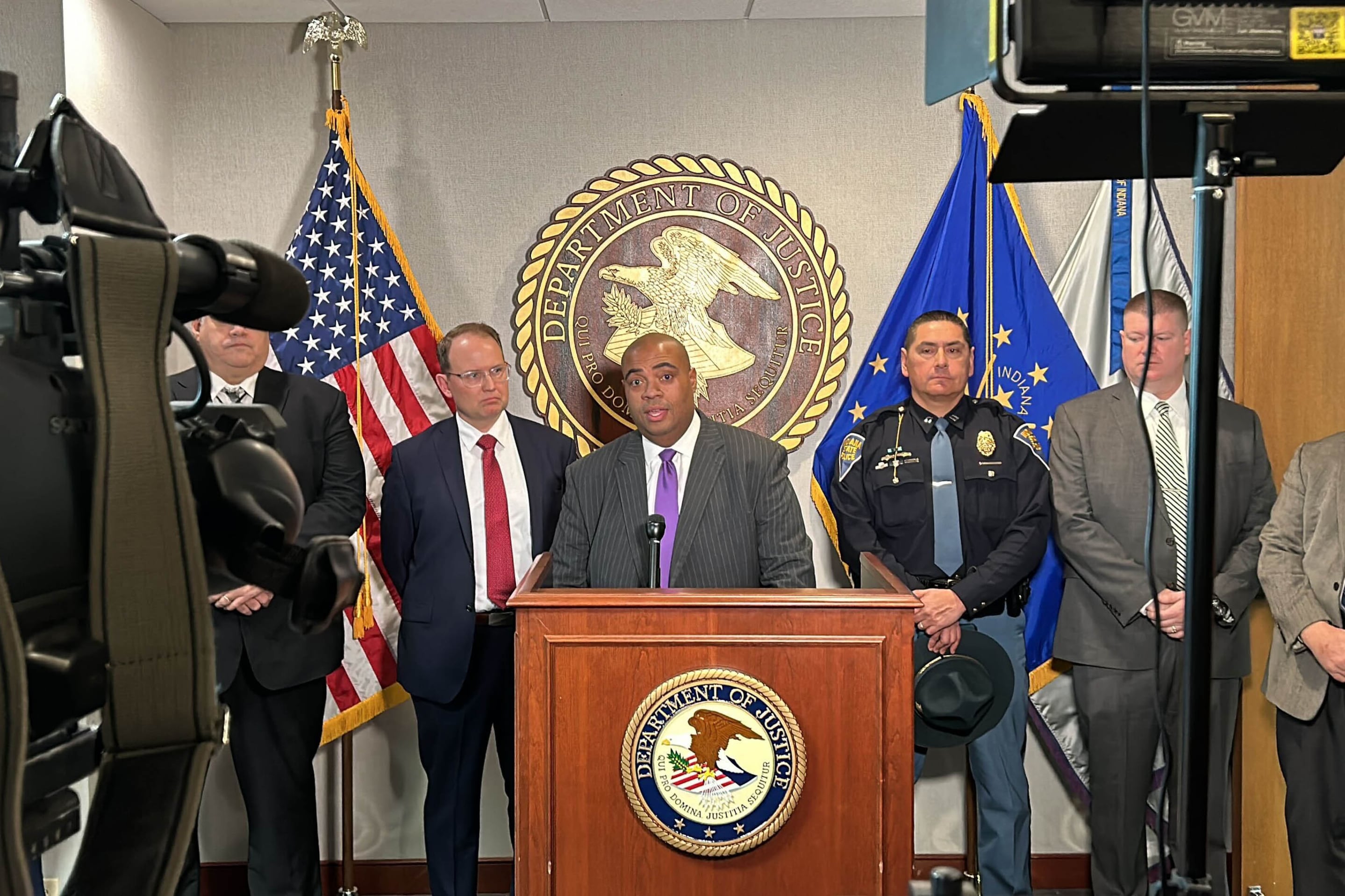 Six men wearing suits stand in a row while the person in the middle stands behind and speaks from a podium. There are two flags and a giant seal in the background and camera gear in the foreground.