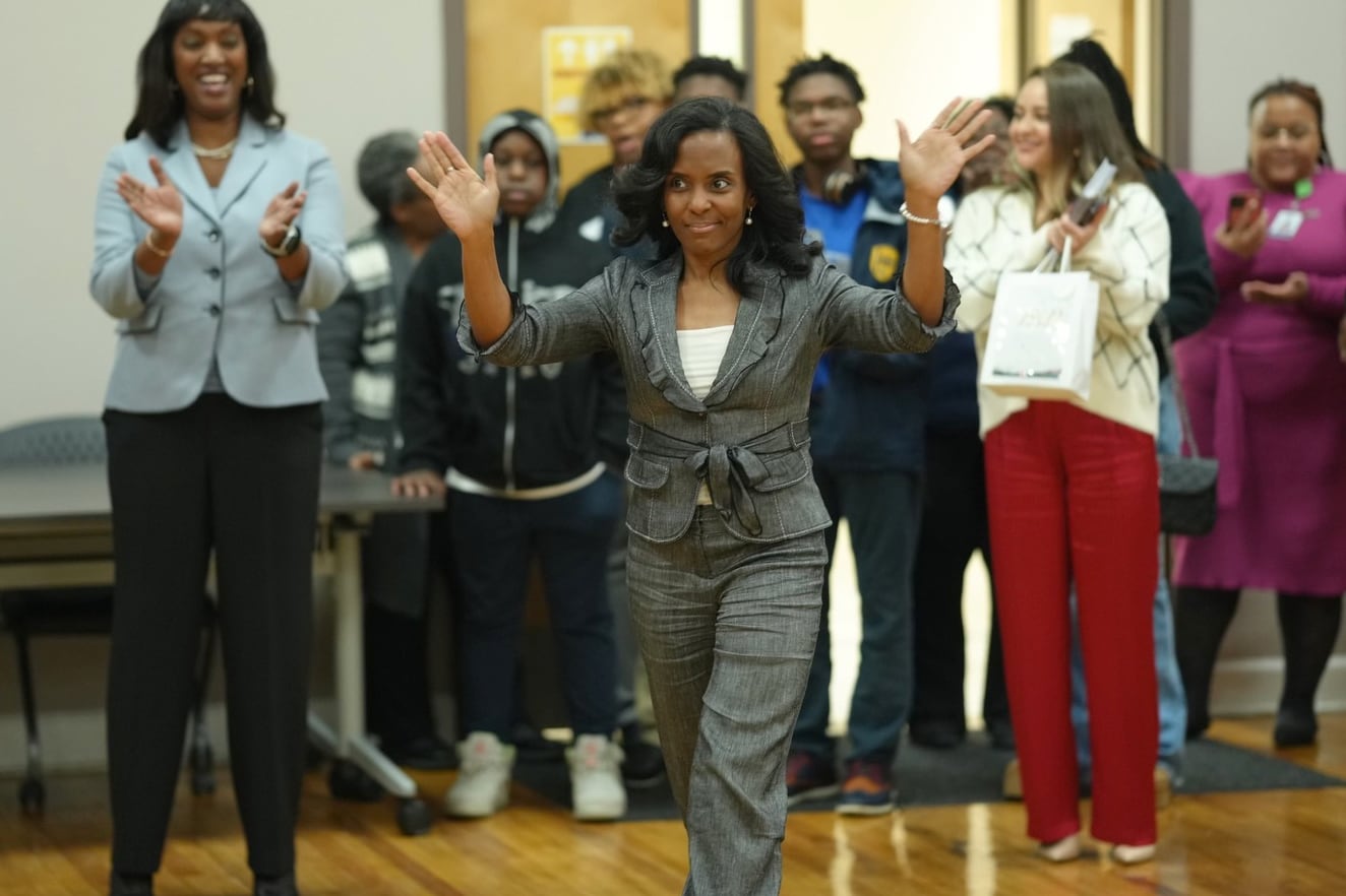 A woman wearing a dark suit jacket waves and walks in front of a crowd of people in the background.