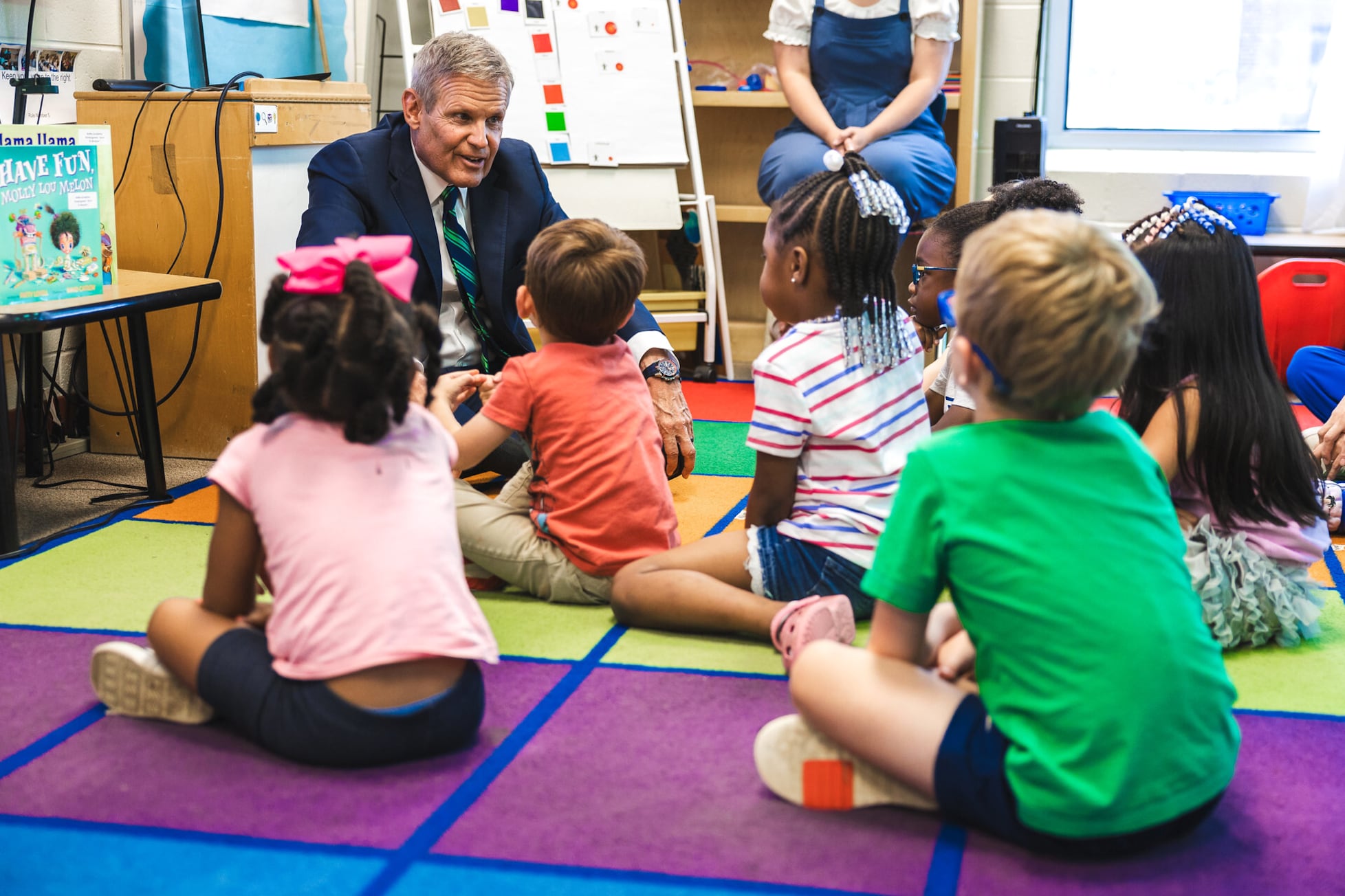 A man in a dark suit sits on a play mat with a bunch of young children in a classroom.