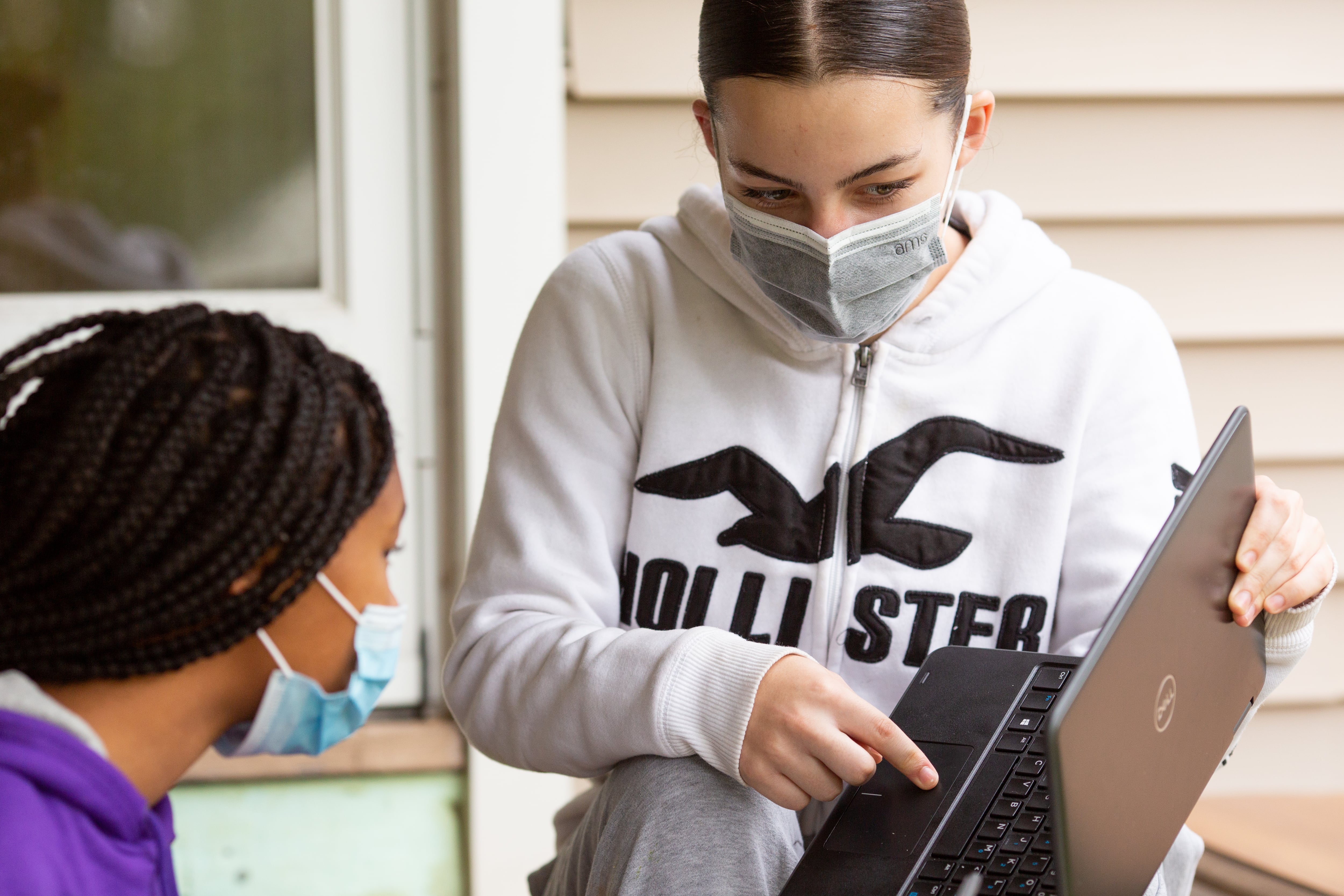 A masked high school student points at a screen on her laptop while another masked student looks on.