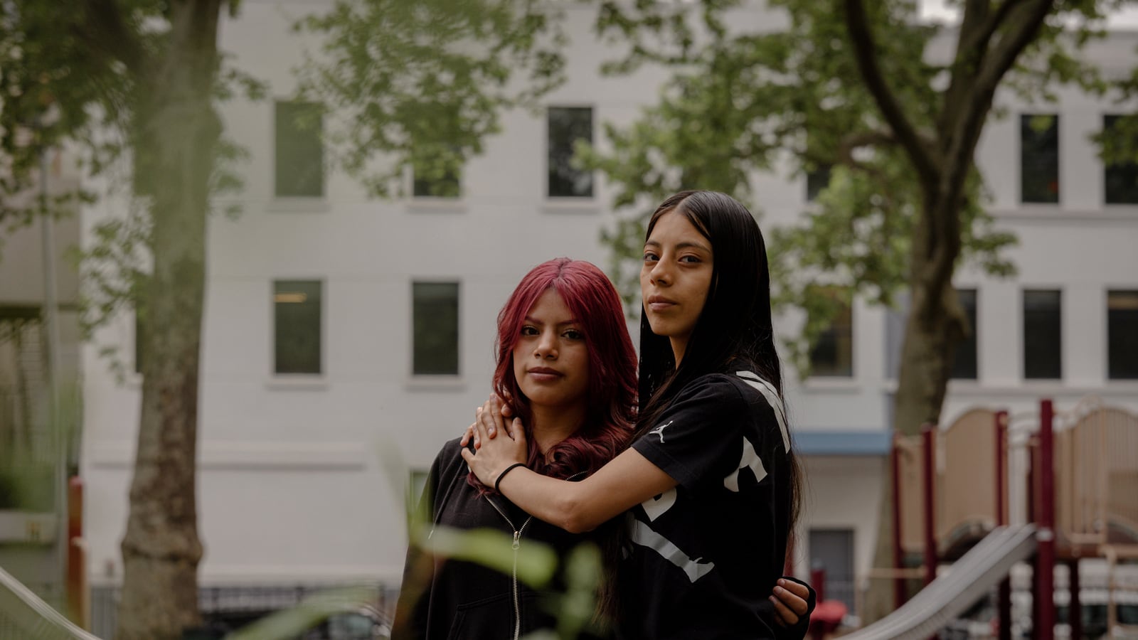 Two high school students and sisters embrace while posing for a portrait in front of a white stone building and a playground.