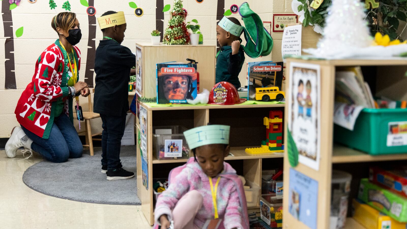 A teacher works with students behind bookcases in a classroom, as a young girl sits on the floor in the foreground.