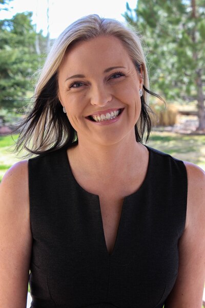 A woman with short blonde hair and wearing a black blouse smiles for a portrait outside with green trees in the background.