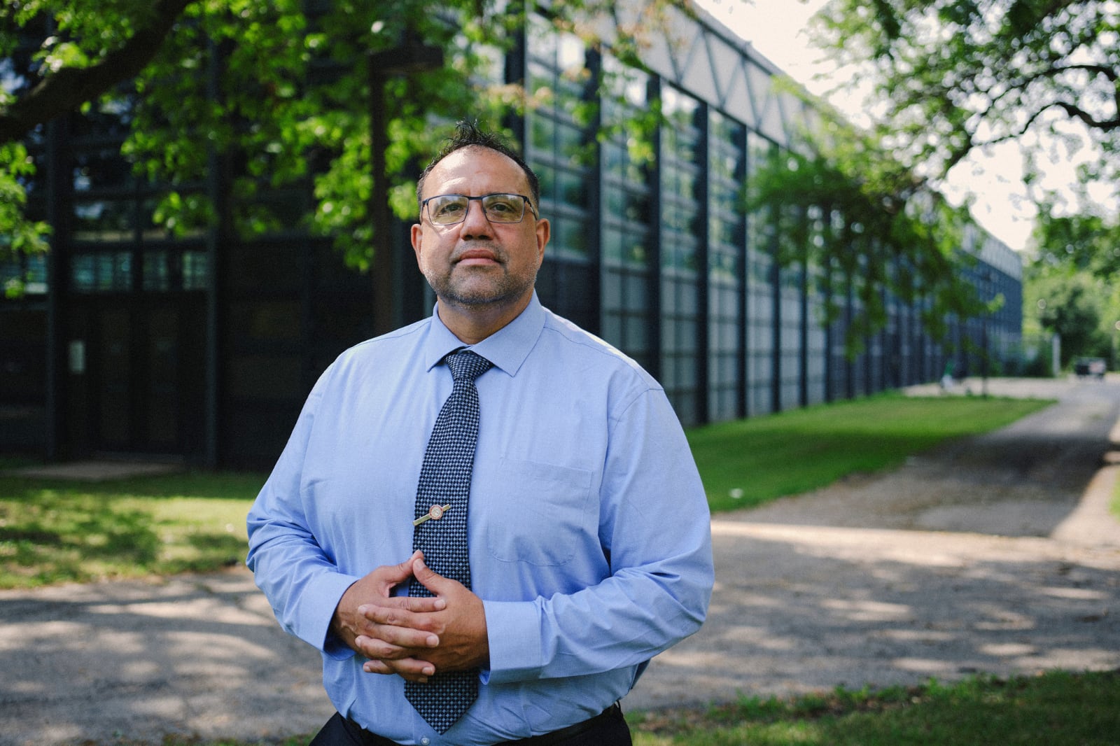 A man in a suit and tie poses for a portrait outside of a large building on a sunny day.