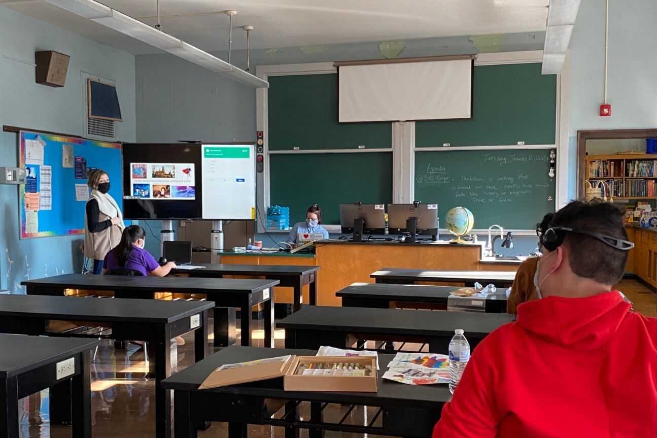A teacher and students several feet apart sit at desks, working in a classroom